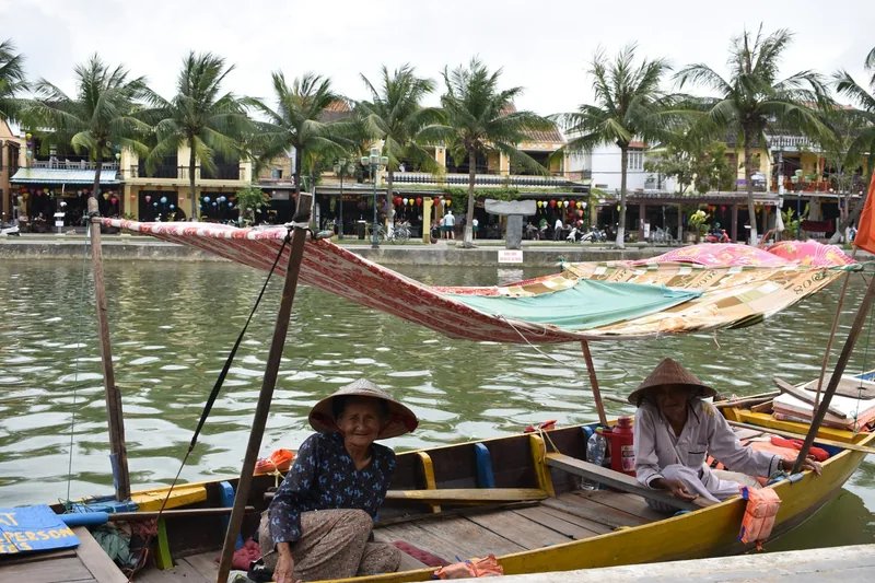 Two boat operators in conical hats sit in wooden boats on the Thu Bon River in Hoi An, with the old town skyline behind.