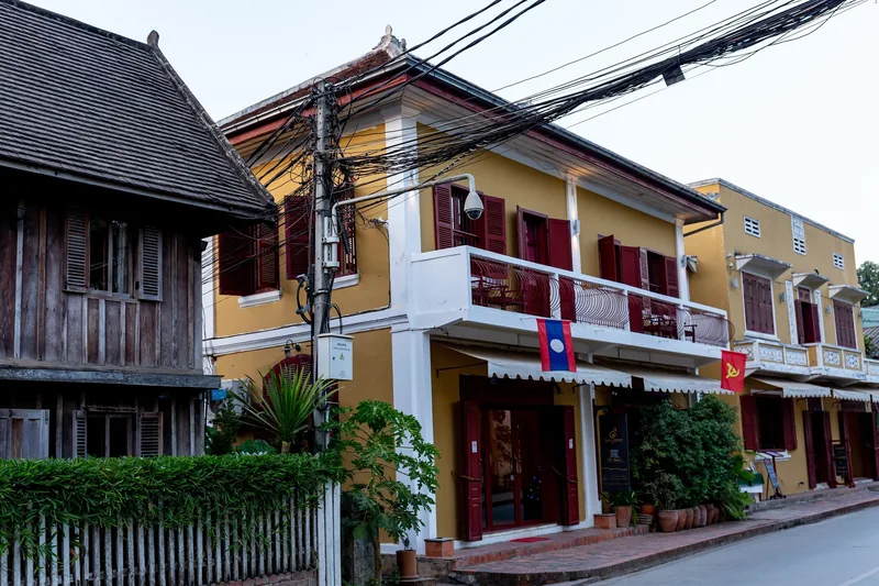 A colonial-era yellow building with red shutters lines a street in Luang Prabang, Laos flags hanging from its balcony.