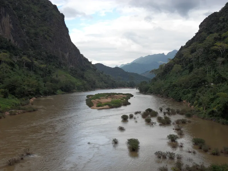 A muddy river winds between steep limestone cliffs near Nong Khiaw, Laos, with a small sandy island dotted with shrubs.