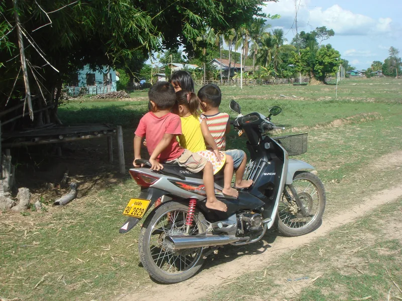 Three children sit on a parked motorbike beside a rural dirt road in Si Phan Don, Laos.