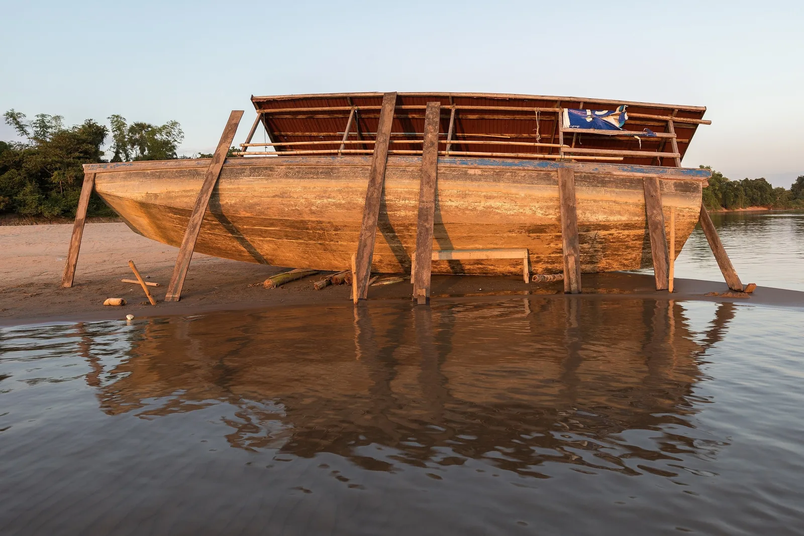 A large wooden pirogue rests propped on stilts along a sandy riverbank in Si Phan Don, Laos.