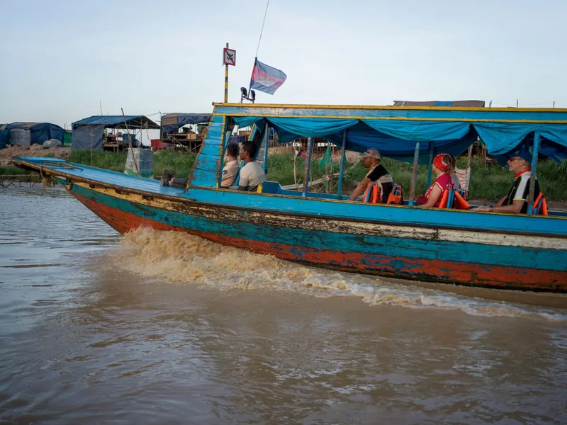 A colorful wooden motorboat carries tourists across muddy river waters near a Cambodian floating village.