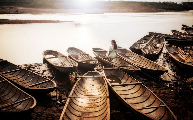 A woman in a light dress sits amid rows of wooden boats on a muddy riverbank in Laos.